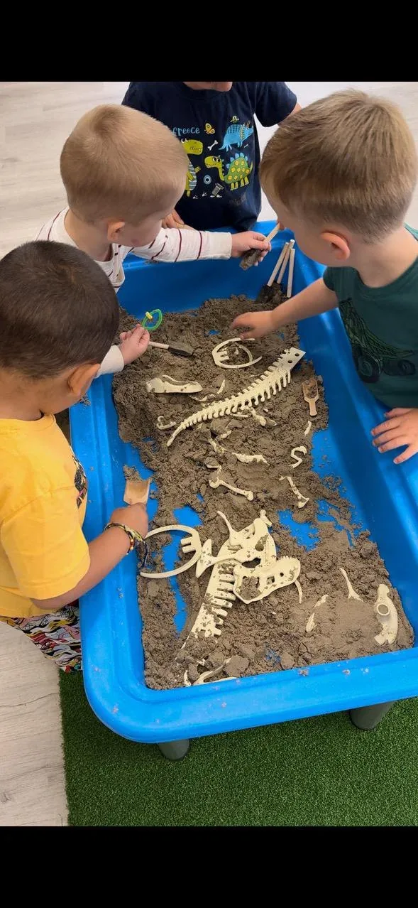 Three children excavating a toy dinosaur skeleton from a sand sensory table