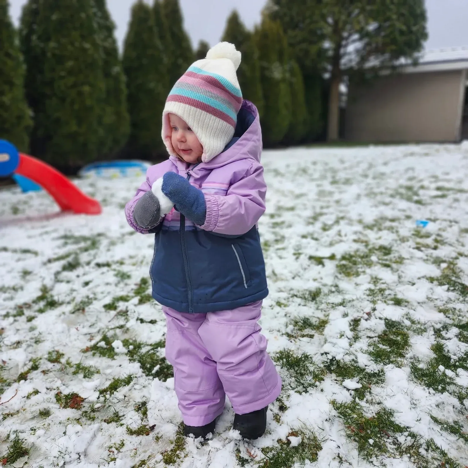 A bundled-up toddler holding a snowball on a snowy playground